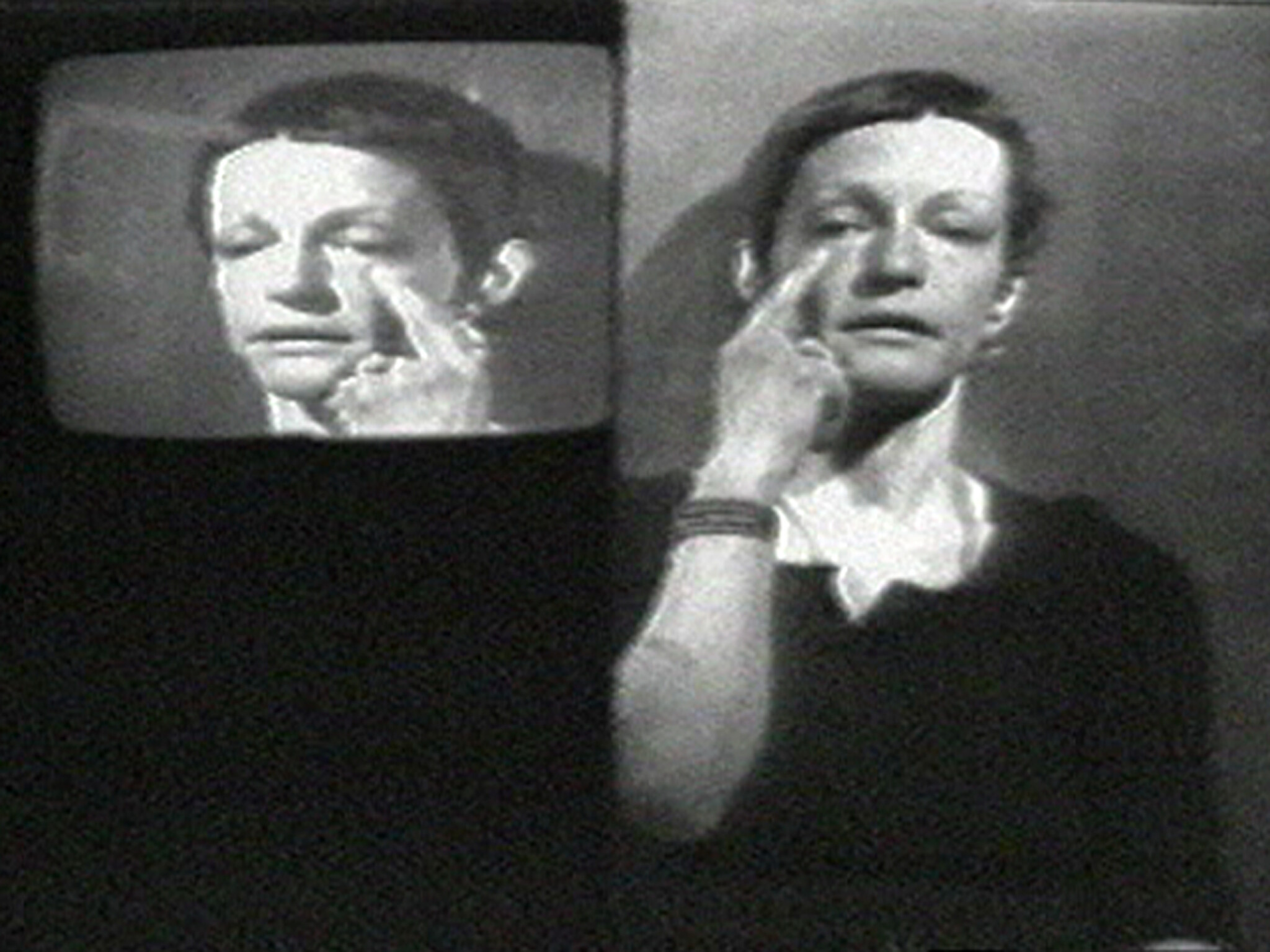 A person touches their face below the eye, with their image also shown on a television screen beside them.