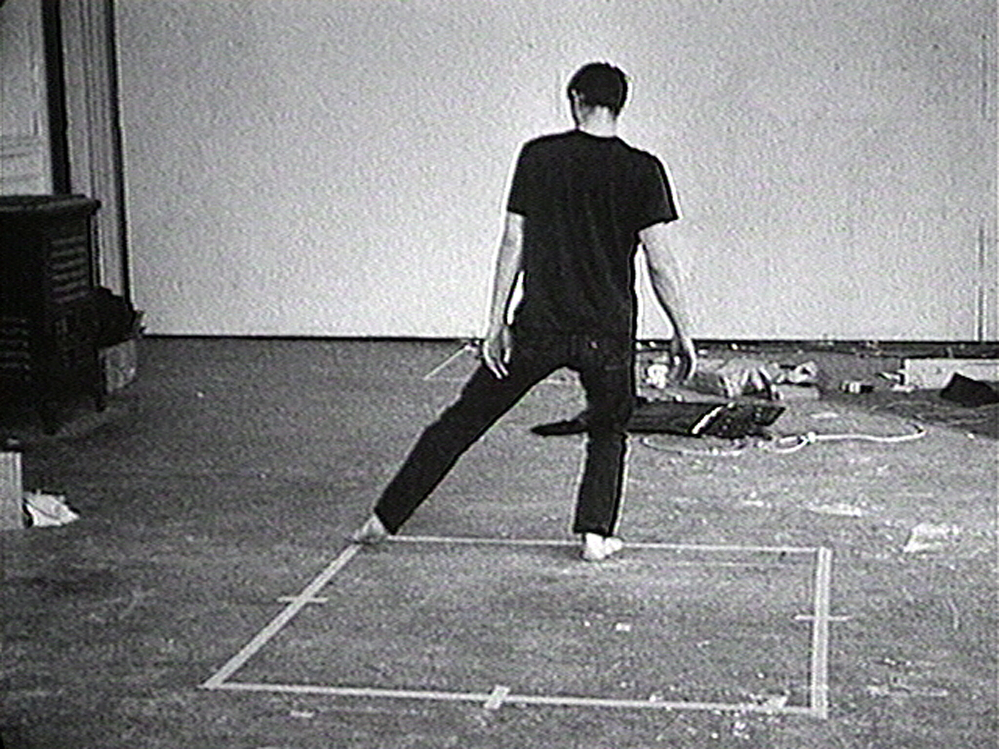 A barefoot person stands inside a taped square on the floor with one leg extended.