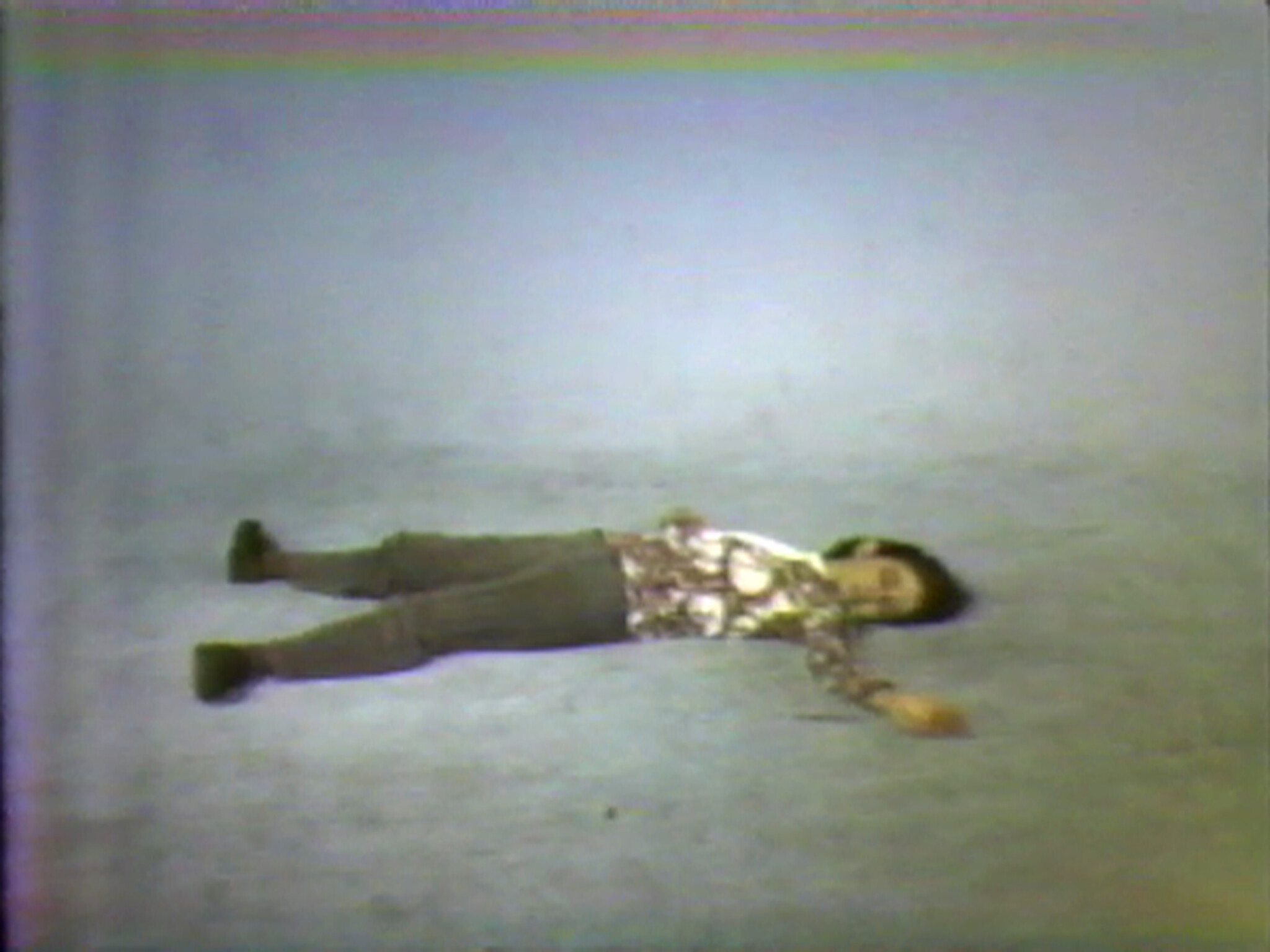 Person wearing patterned shirt lies face up on a plain floor with one arm extended.