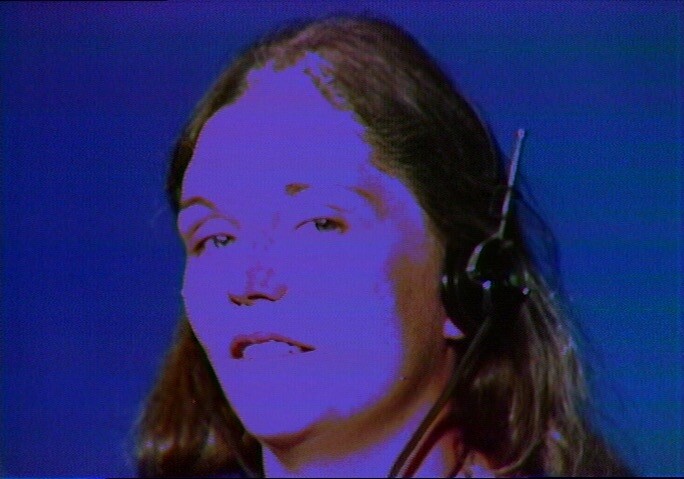A person with long hair wears a headset microphone and looks forward against a blue background.