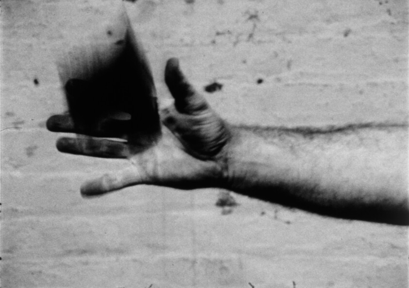 A hand is tossing a small dark object into the air against a plain background.
