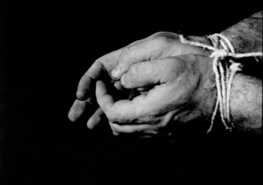 Two hands bound at the wrists with rough rope against a dark background.