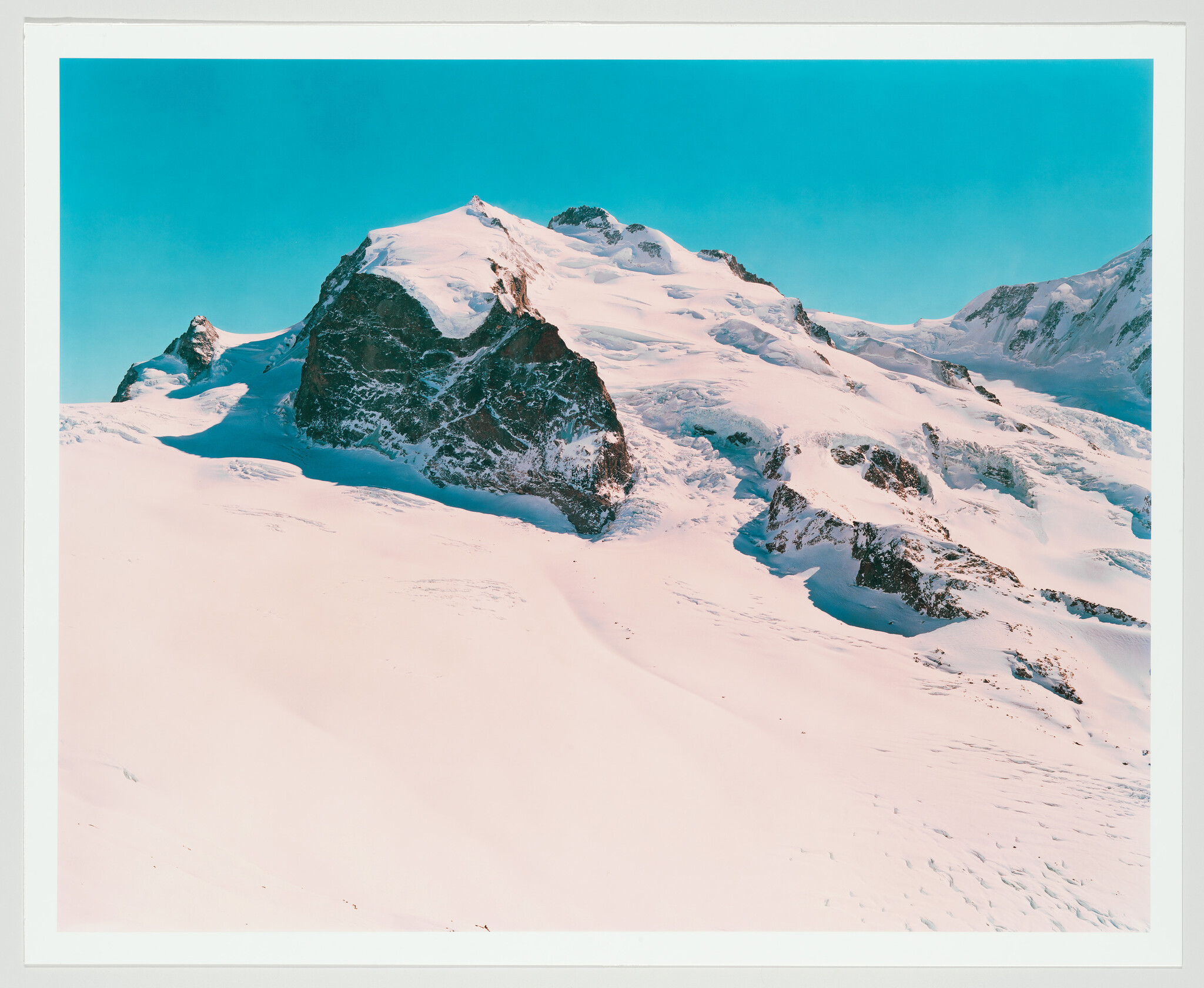 Snow-covered mountain peaks rise above a wide snowy slope under a clear blue sky.