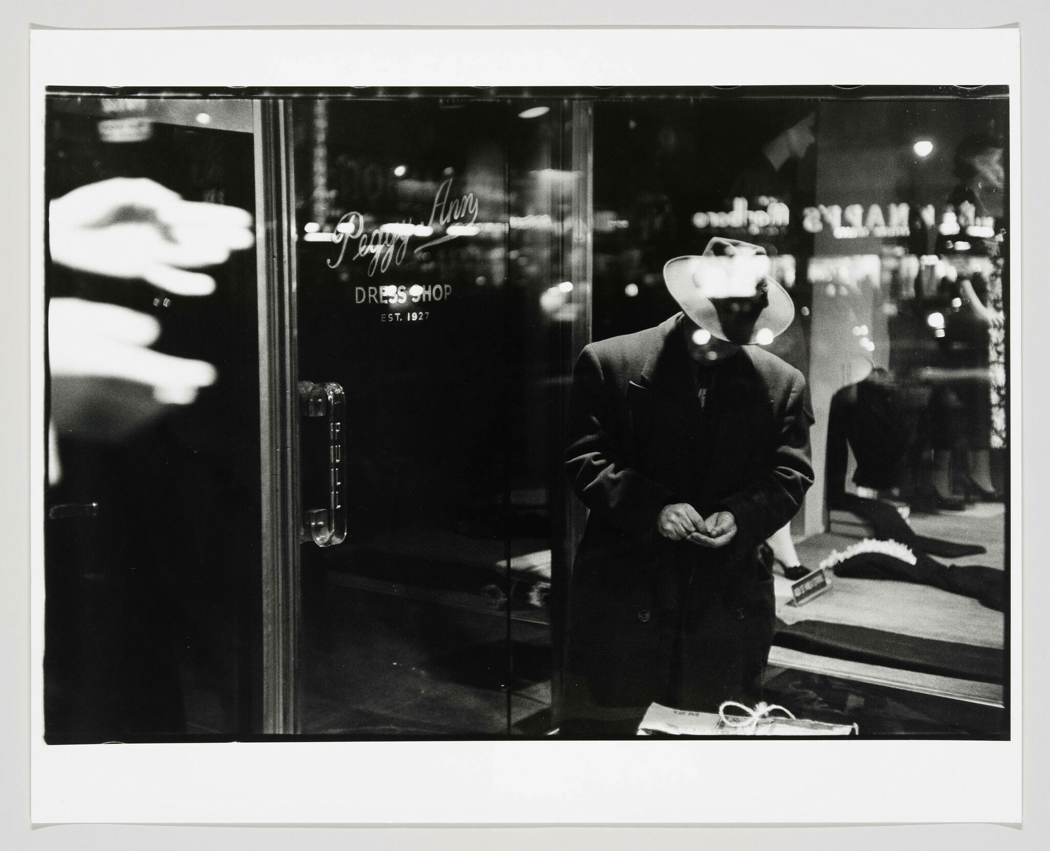 Black and white photograph of a mannequin dressed in a dark coat and a wide-brimmed hat, standing behind a glass door of a dress shop named "Peggy Ann." The reflection of neon signs and the street scene is visible on the glass, creating a layered visual effect. A newspaper and a pair of gloves rest on a ledge in front of the mannequin.