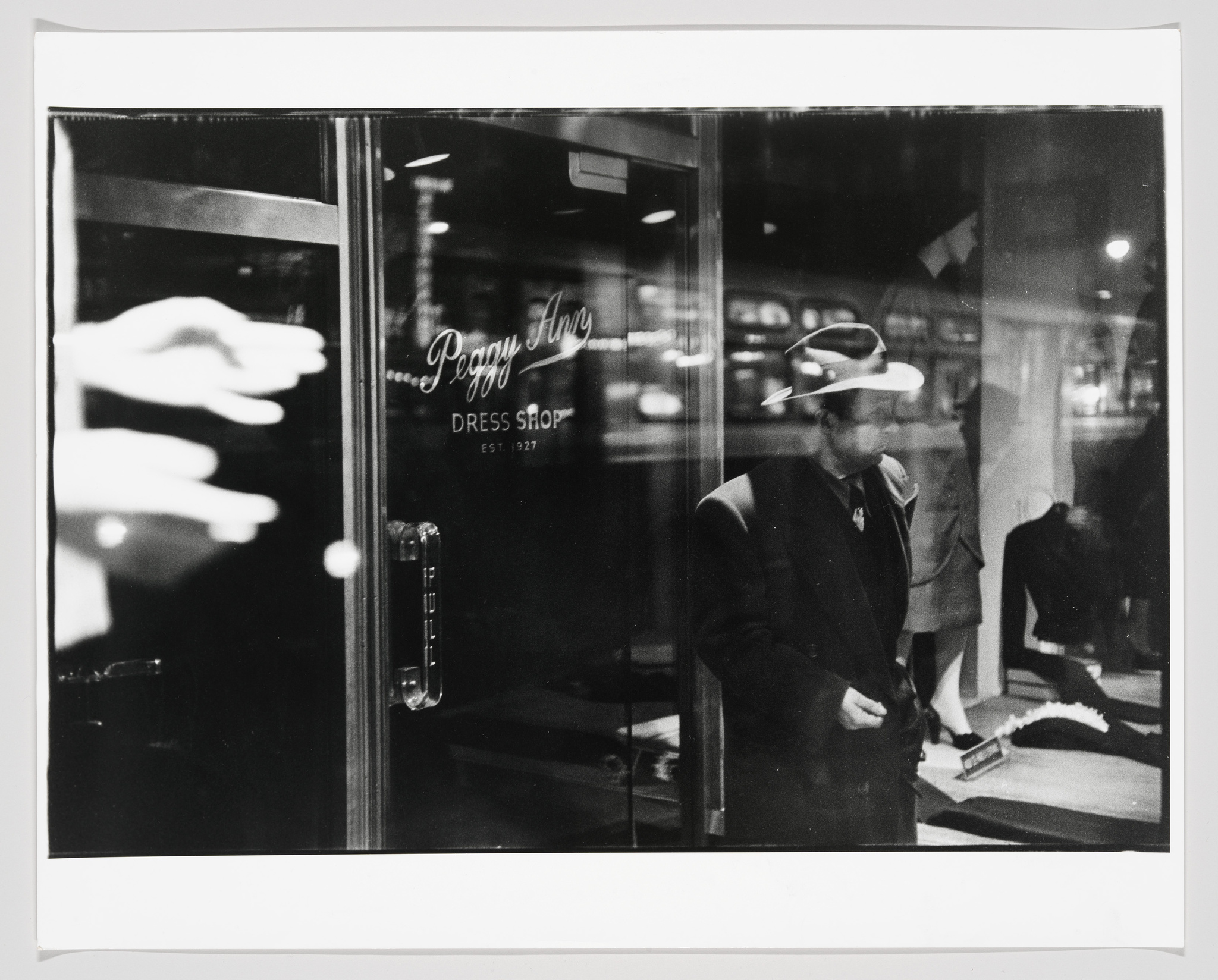 A black and white photograph capturing a reflective glass door of the "Peggy Ann Dress Shop" with a man in a fedora hat visible through the glass, seemingly standing inside the shop. The reflection shows a blurred hand pressing against the glass and city lights, creating a layered, ghostly effect.