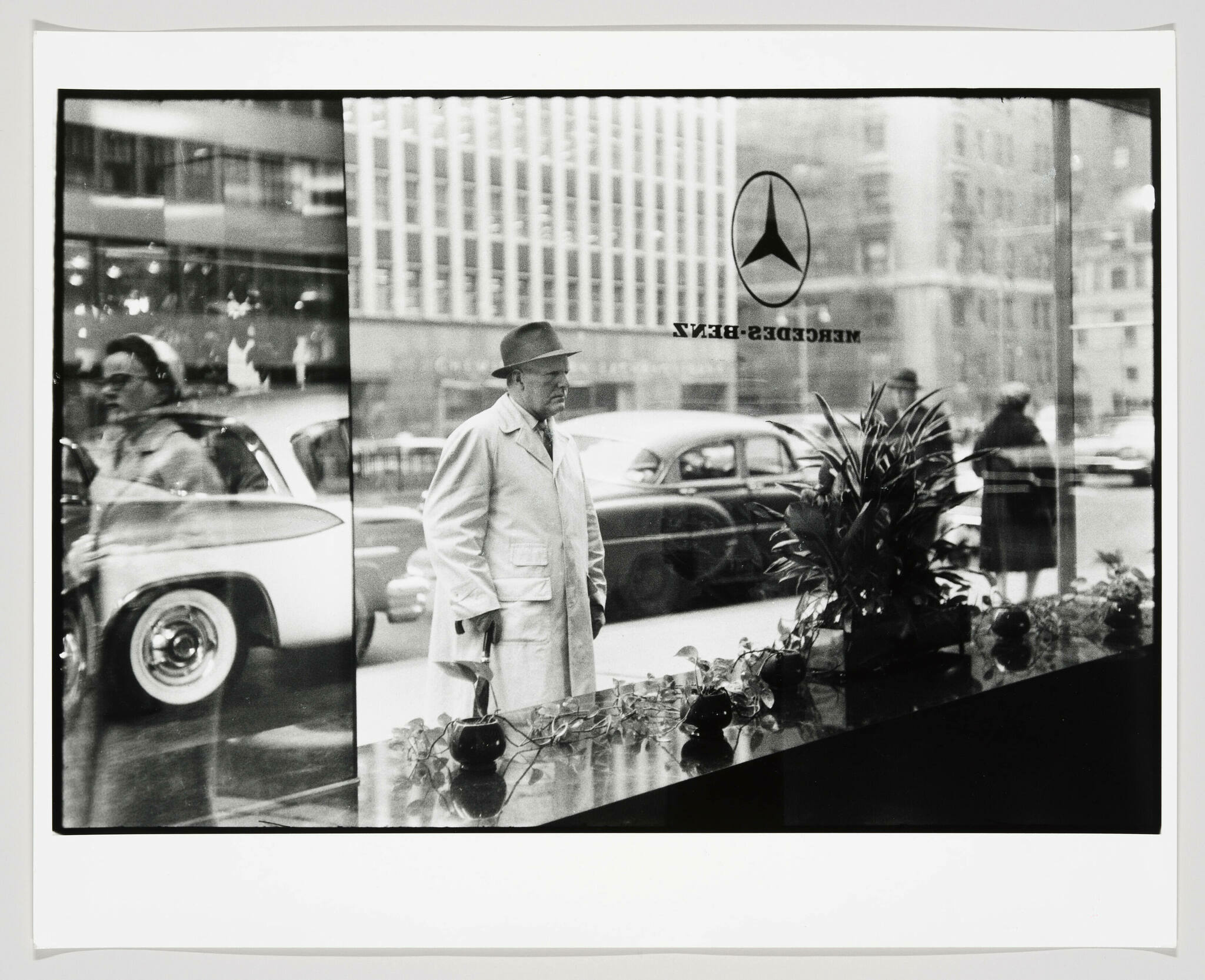 Black and white photograph capturing a street scene through a window with reflections. A man in a trench coat and hat stands to the right, looking to the side, with the Mercedes-Benz logo and reversed lettering "MERCEDES-BENZ" visible on the window. The reflections show cars and pedestrians on the busy street outside.