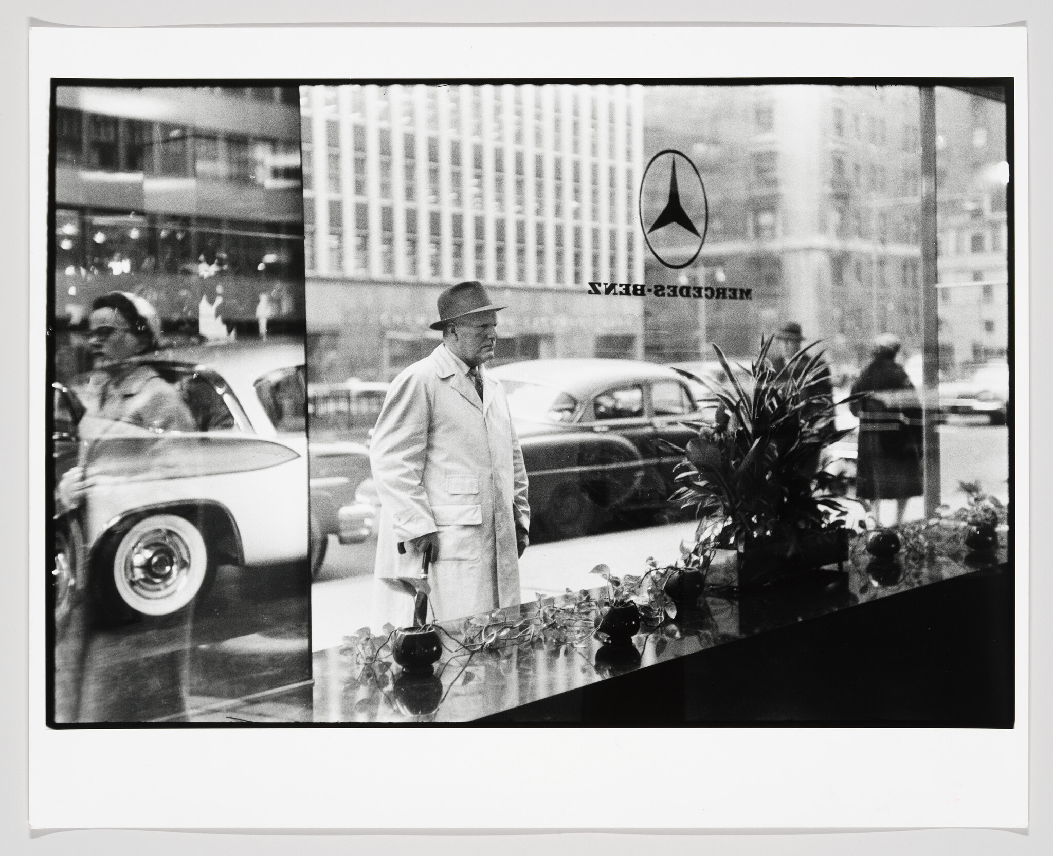 A man in a coat and hat stands outside a Mercedes-Benz showroom window, looking in at the display.