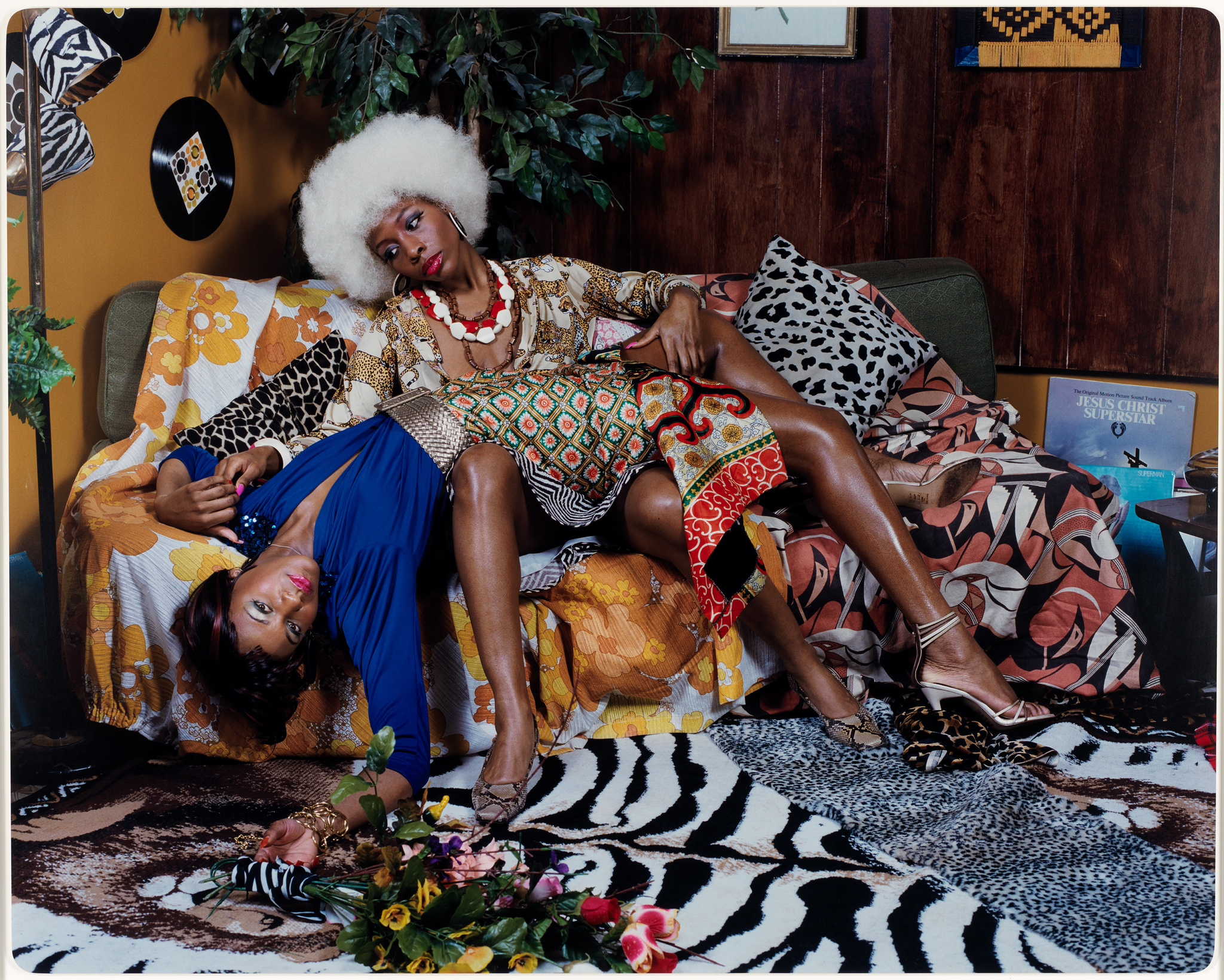 Two Black women in richly-patterned clothing sprawl on a sofa, surrounded by artificial foliage and animal-print textiles
