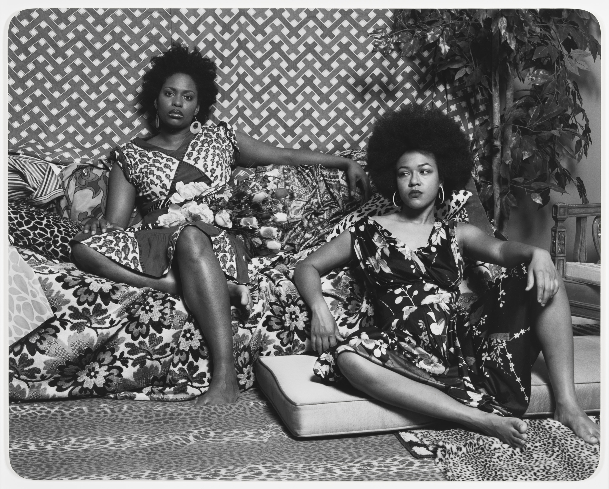 Two Black women wearing vibrantly patterned dresses sit in a similarly patterned interior.