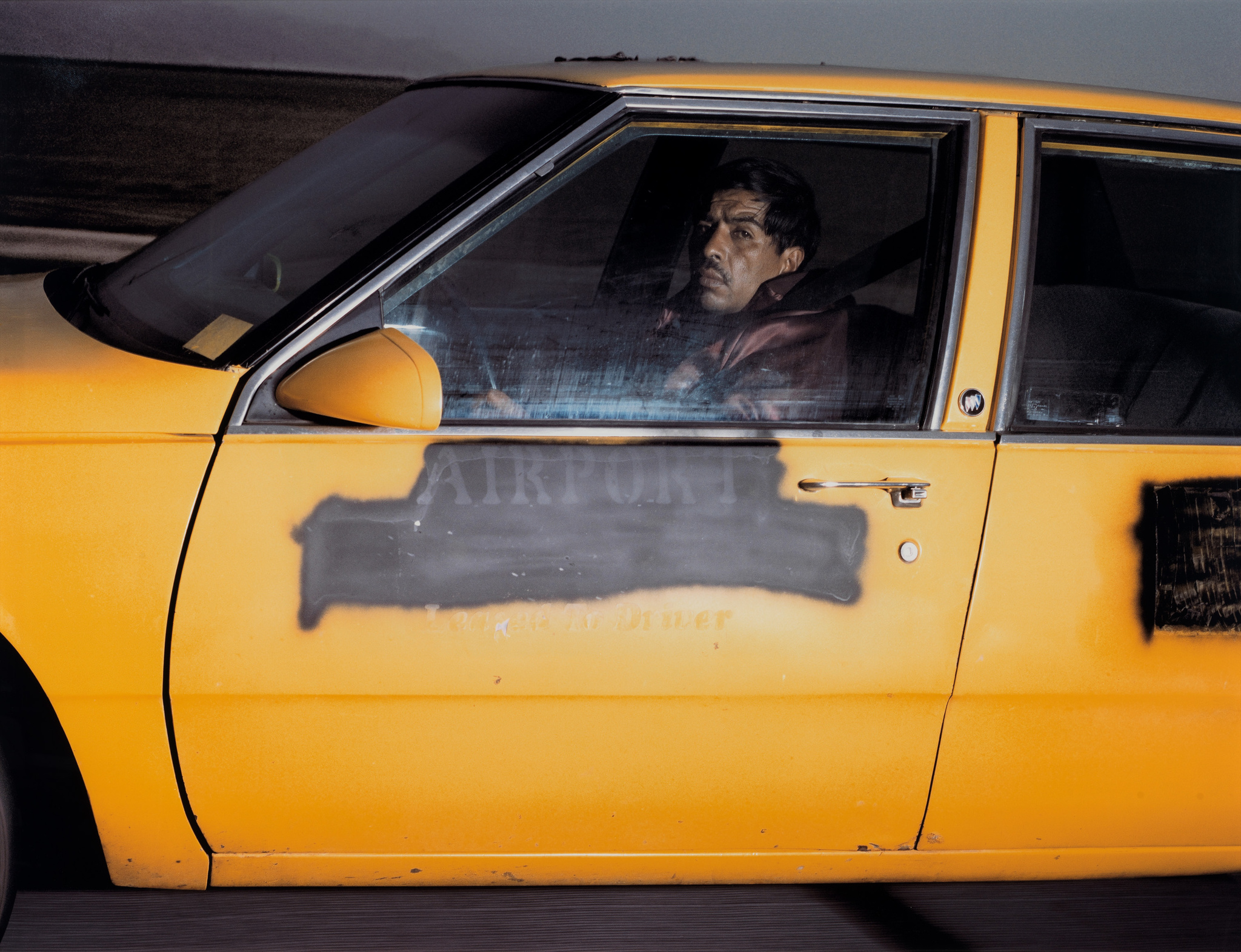 A man is seen through the window of a moving yellow taxi cab, with the word "AIRPORT" reflected on the door, suggesting the vehicle is in transit to or from an airport. The man appears to be the driver, looking intently ahead, possibly focused on the road or traffic. The image captures a moment of everyday life, possibly in an urban setting.