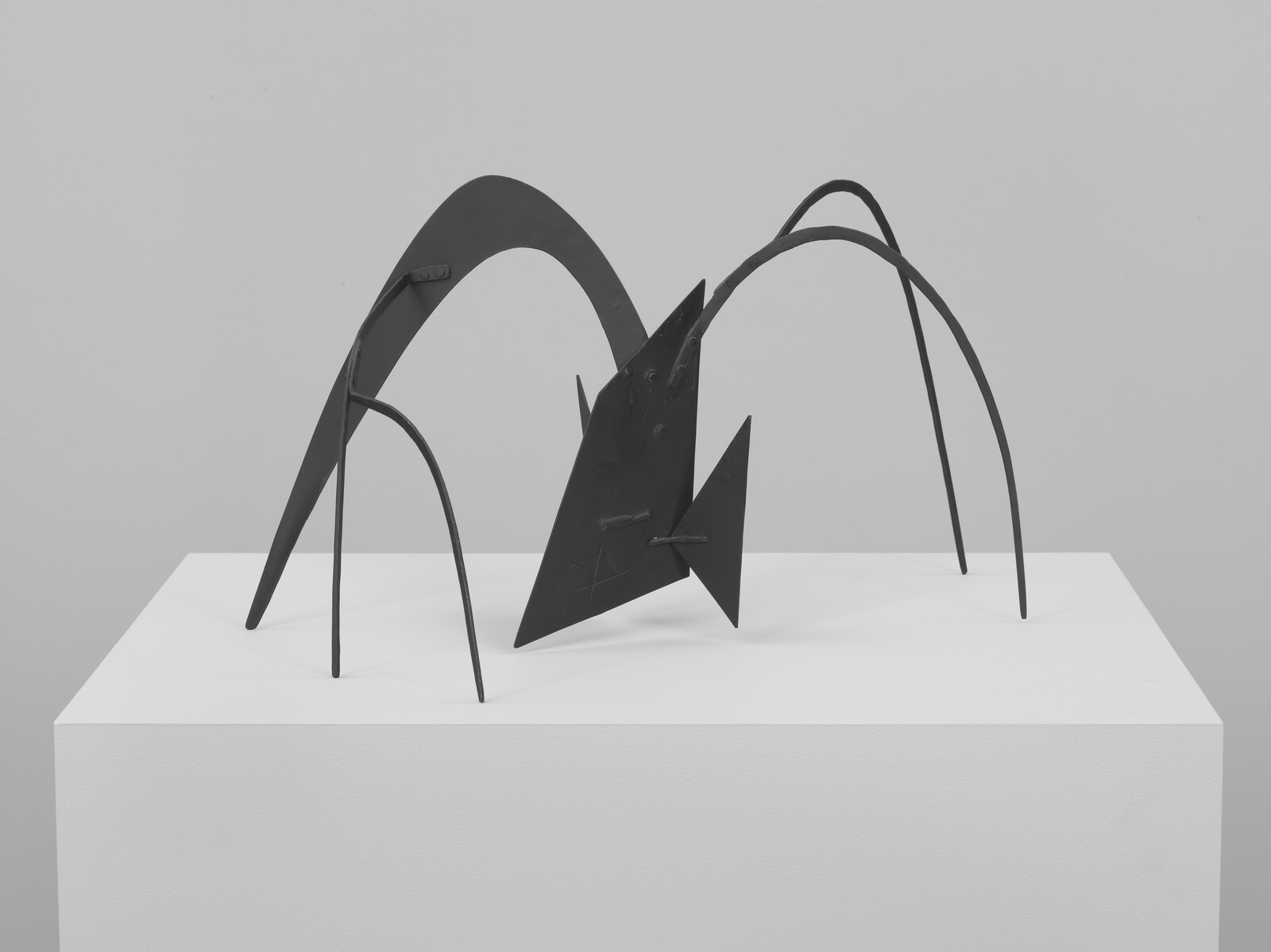 Abstract black metal sculpture with arched legs standing on a white pedestal.