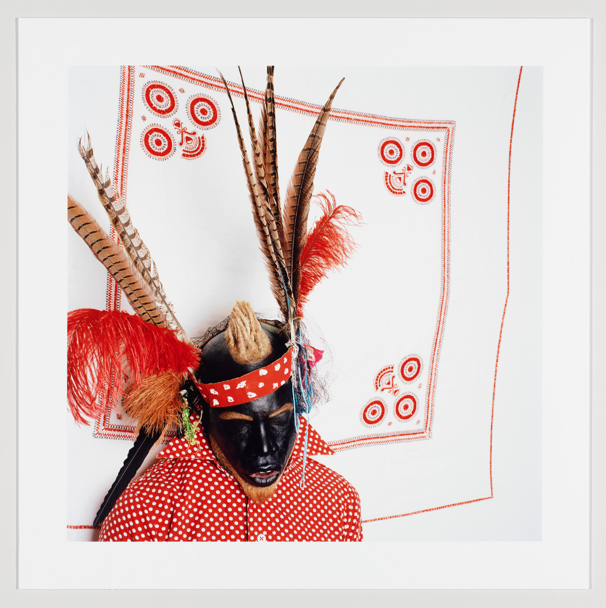 Person wearing black face paint, red polka-dot shirt, and tall feathered headdress facing downward.