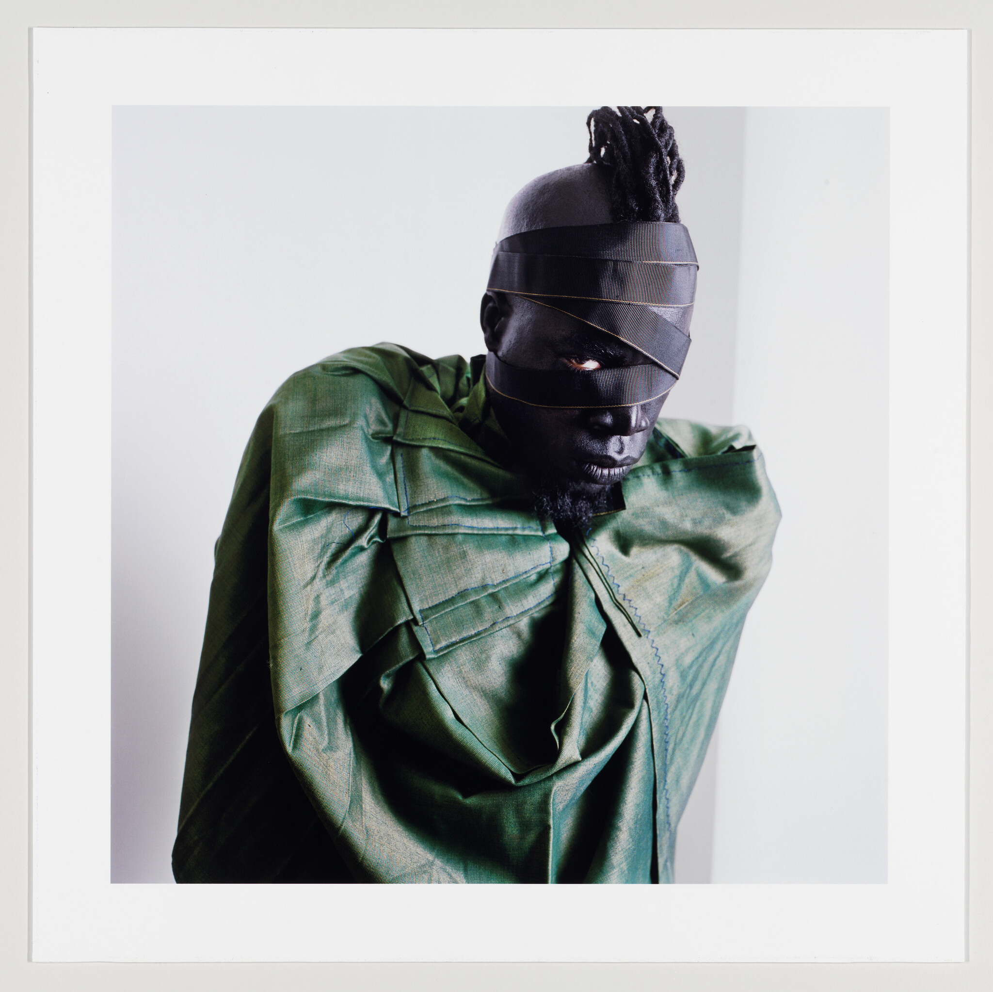 Man with black facial bands and green draped fabric staring with one visible eye.