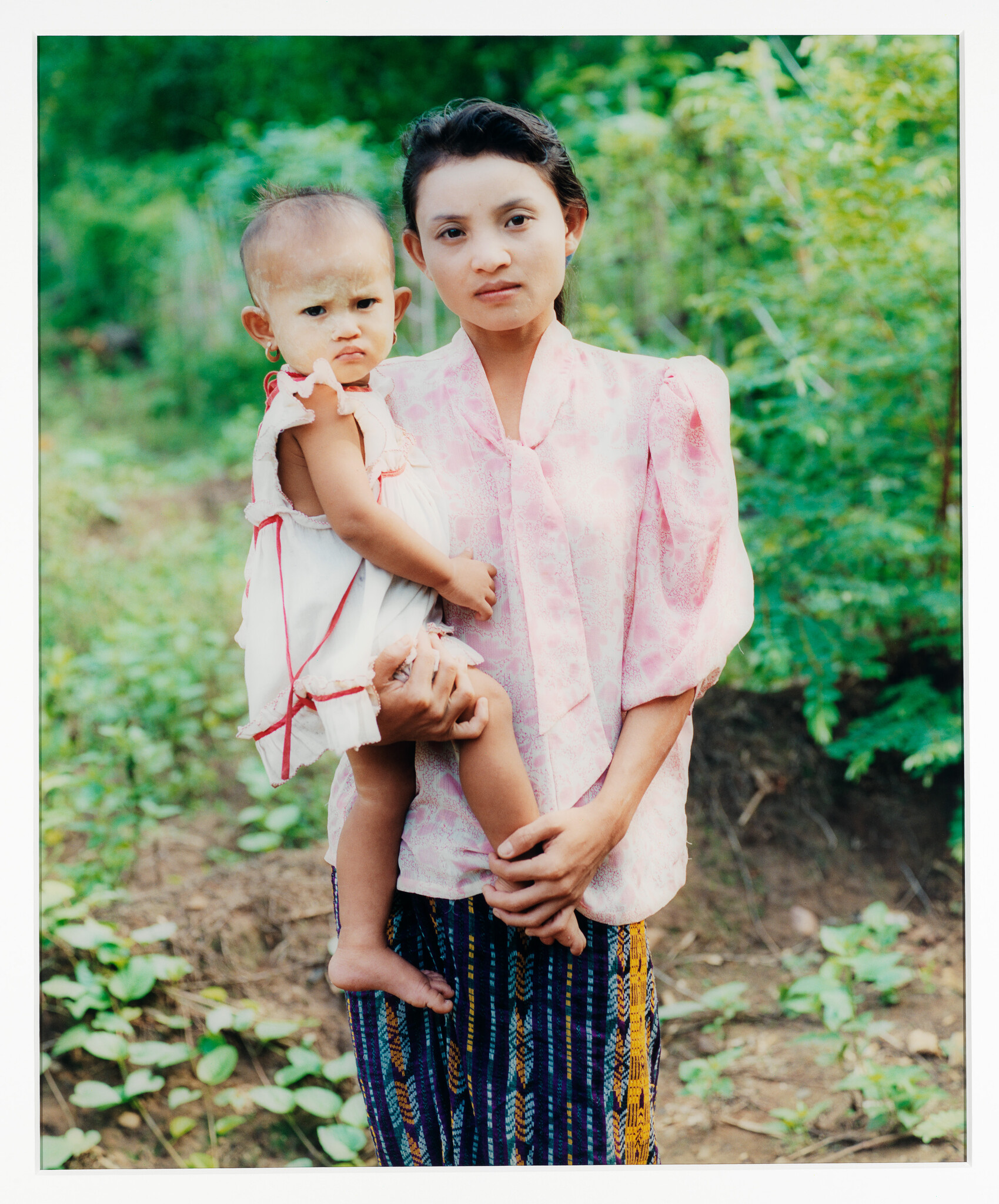 A young woman stands outdoors holding a small child against a green background.