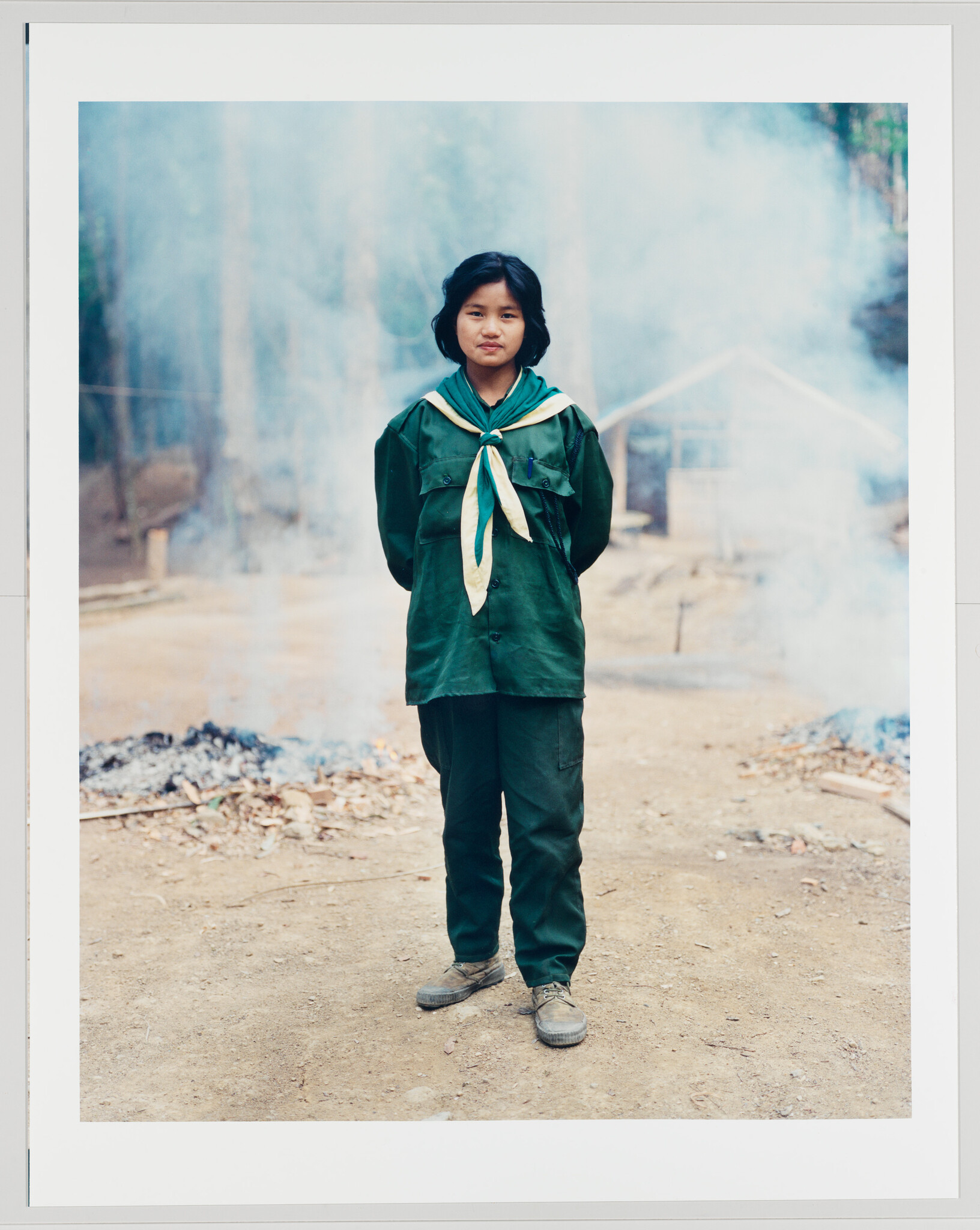 Young girl in a green scout uniform stands on a dirt path with smoke behind her.