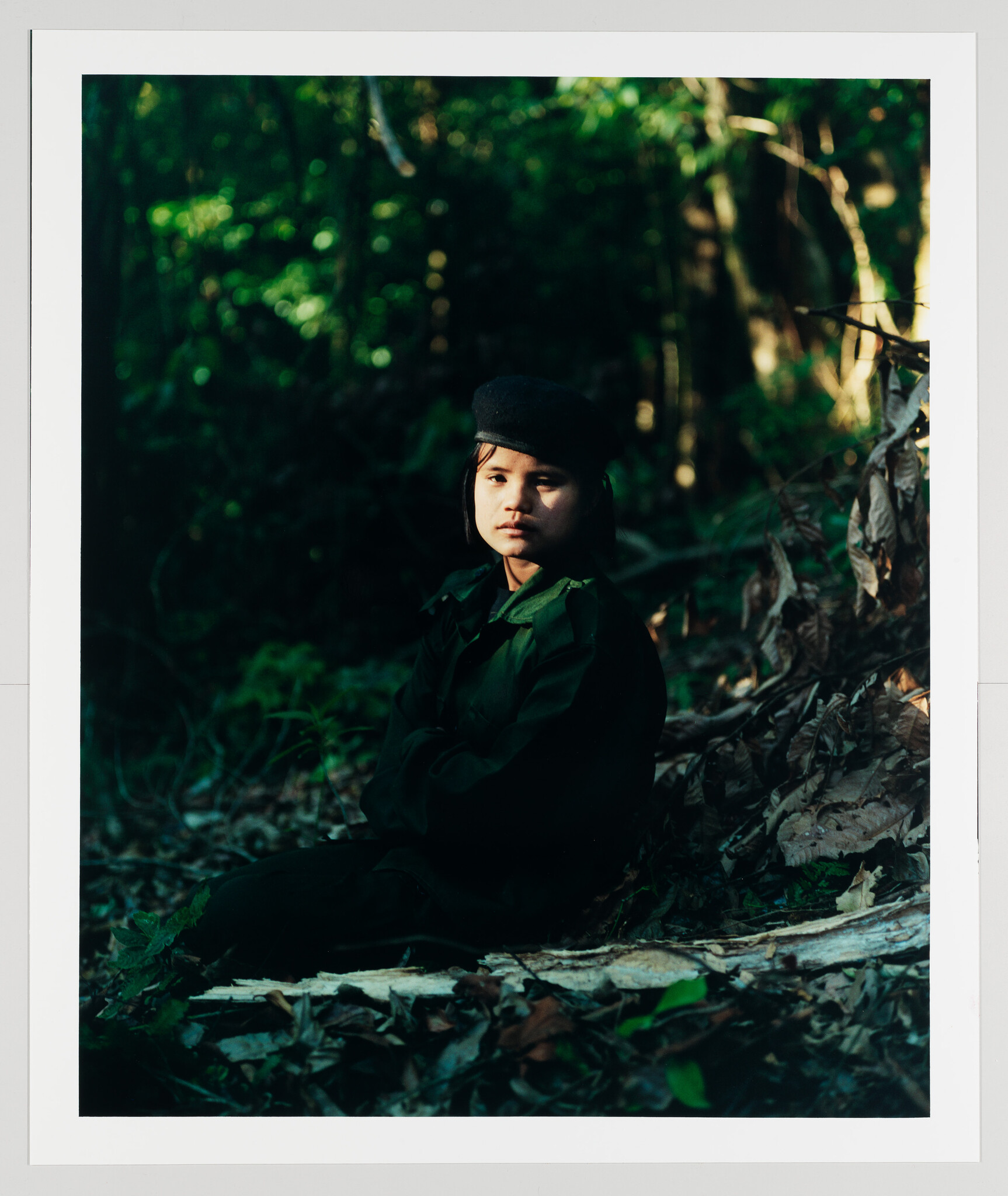 A child in a dark green uniform and beret sits on the forest floor with crossed arms.