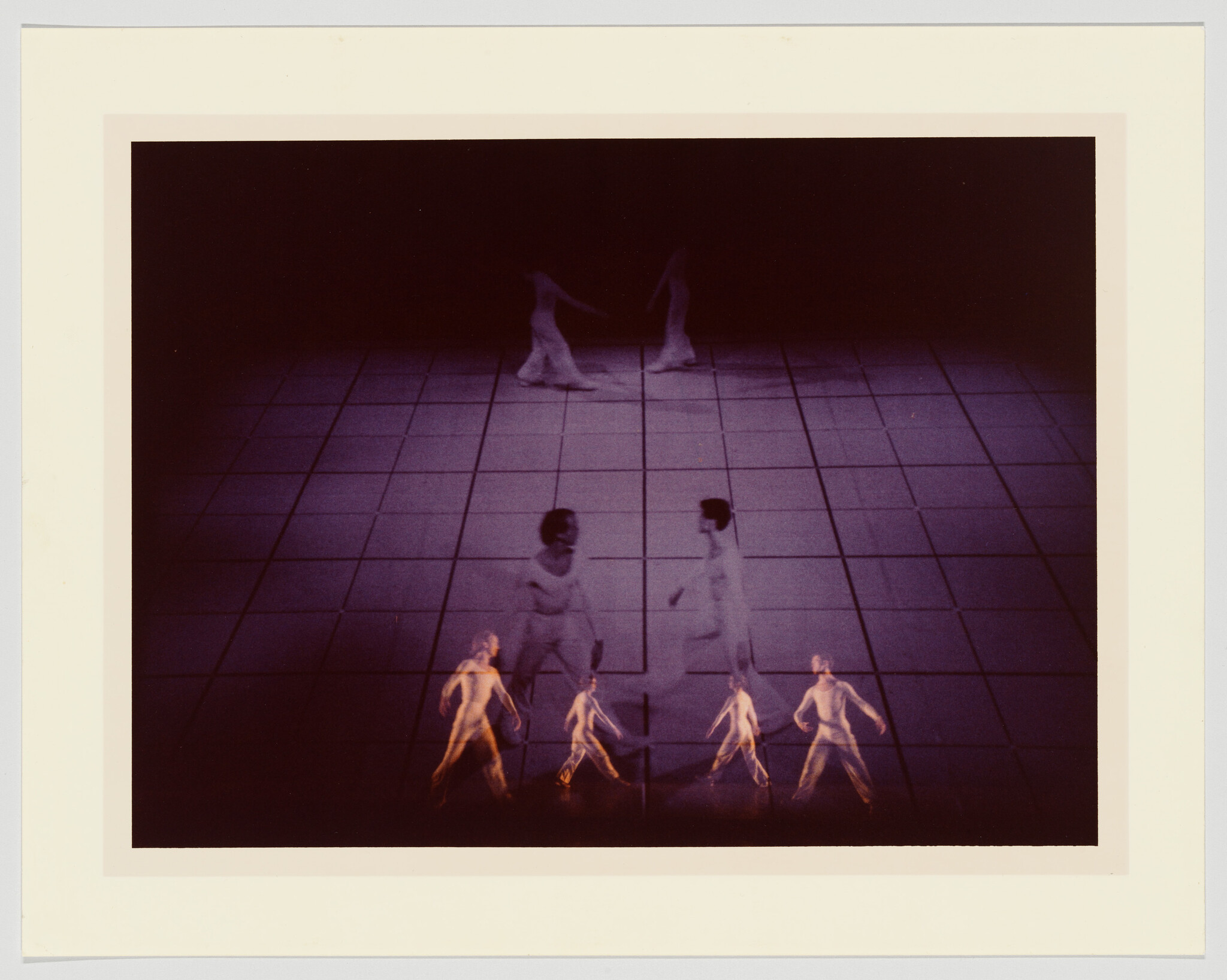 A photograph capturing a dance performance with multiple dancers blurred in motion on a checkered floor, suggesting movement and the fluidity of the dance. The lighting casts a warm hue over the scene, and the dancers appear to be wearing light-colored costumes. The image is framed with a white border.