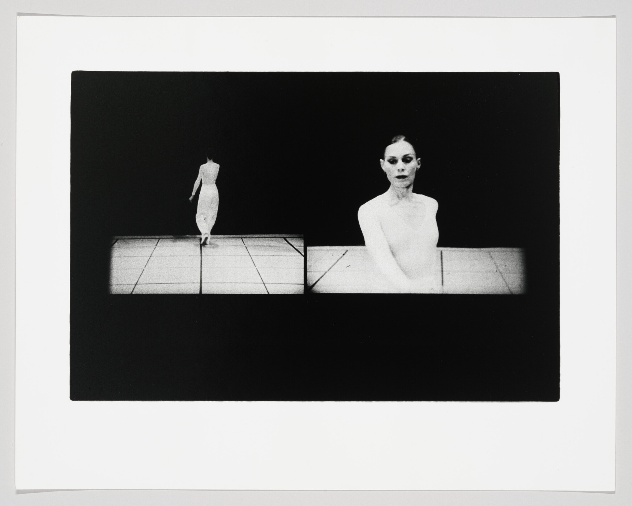 A black and white photograph featuring two figures against a dark background. On the left, a person is seen from behind, walking away on a tiled platform. On the right, another figure is seated facing forward, with a strikingly pale complexion and dark eye makeup, creating a stark contrast with the surrounding darkness. The image has a border, suggesting it is a print.