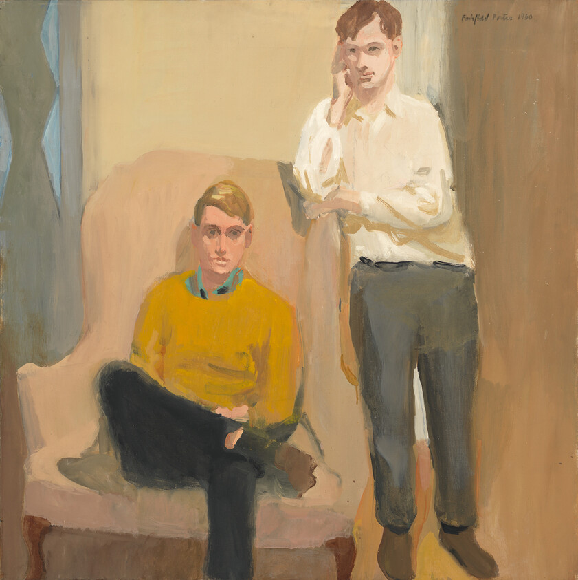 Two young men posed in a room, one seated in a yellow sweater and one standing.