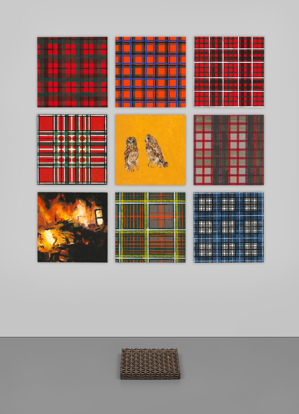 A gallery wall featuring a collection of nine abstract paintings with various patterns and colors, including red and black grids, a yellow canvas with two owls, and a dark painting with fiery tones. A woven brown mat lies on the floor in front of the wall.