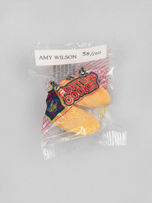 A clear plastic-wrapped pair of fortune cookies in branded packaging with a white label reading a handwritten name and number.