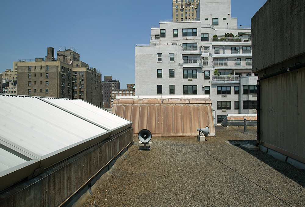 Flat rooftop with gravel surface, two large loudspeakers, and surrounding tall apartment buildings under a clear sky.
