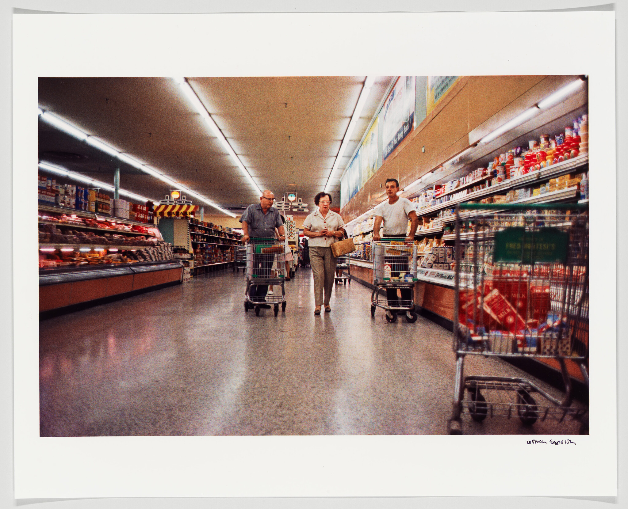 Three shoppers walk down a supermarket aisle pushing shopping carts and talking.