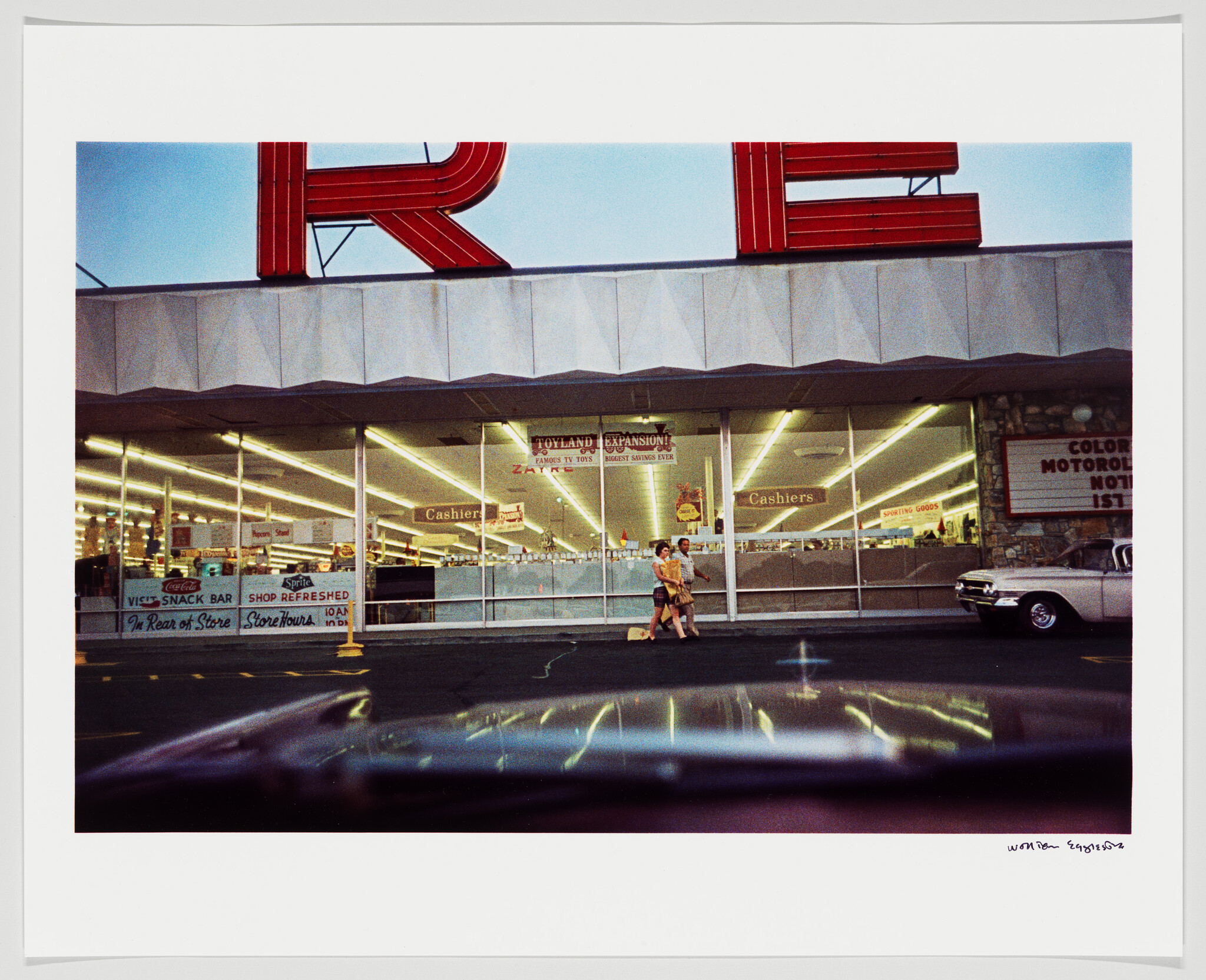 Two shoppers walk past a brightly lit supermarket storefront carrying bags while cars sit in the parking lot.