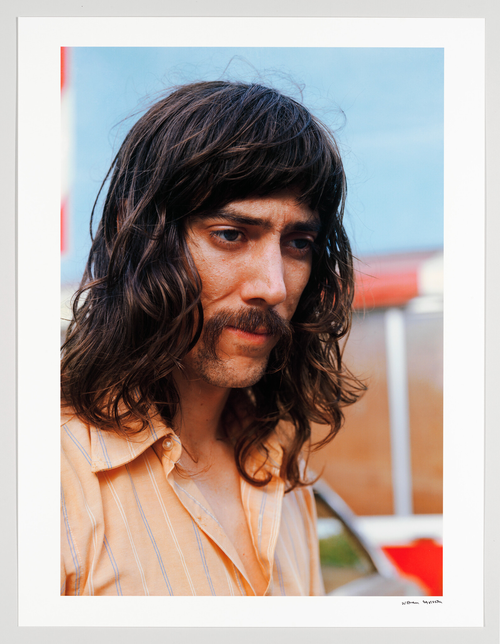 Man with long hair and mustache looks down pensively while wearing an orange striped shirt.