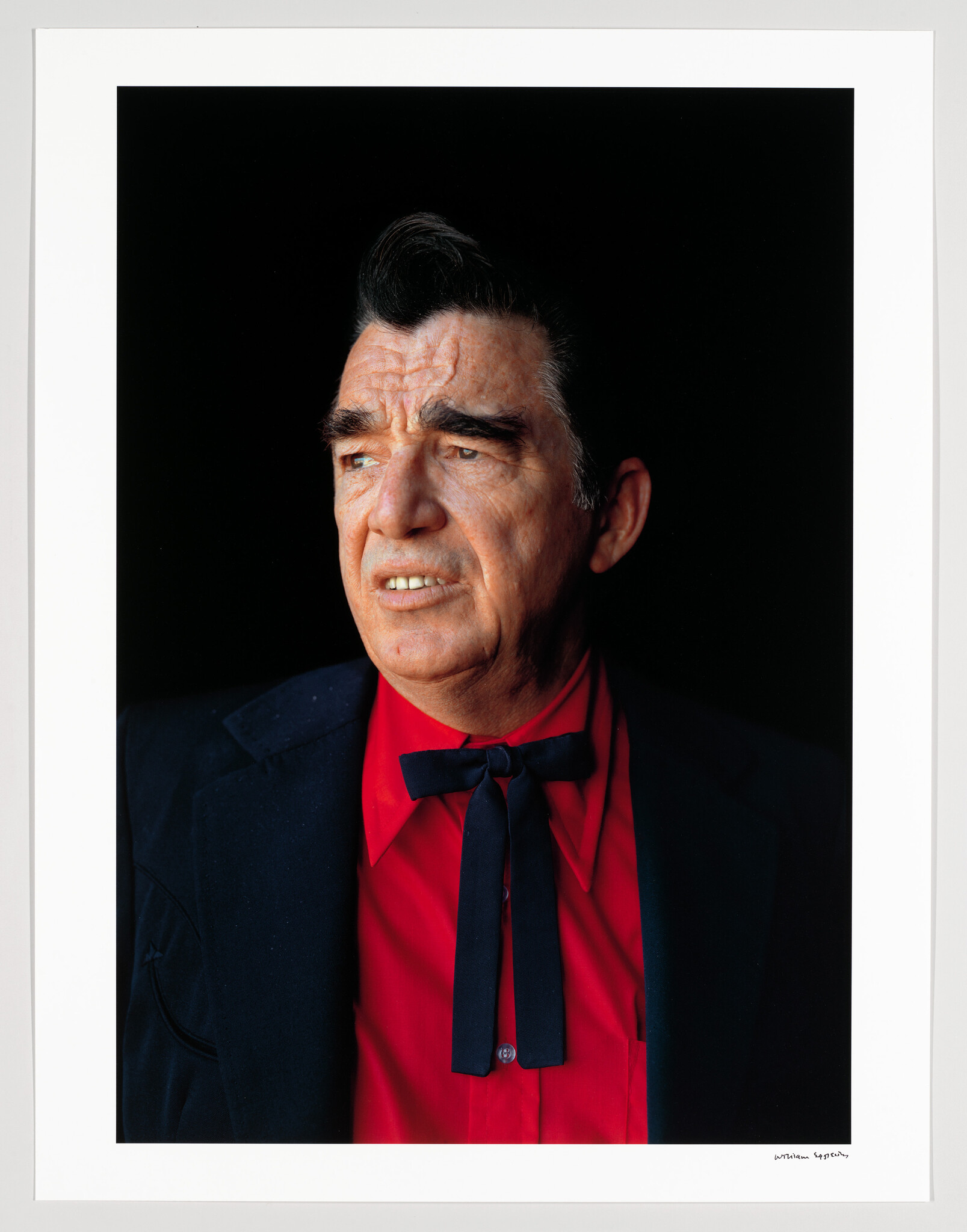 Older man in a red shirt and black bow tie looking thoughtfully to the side.