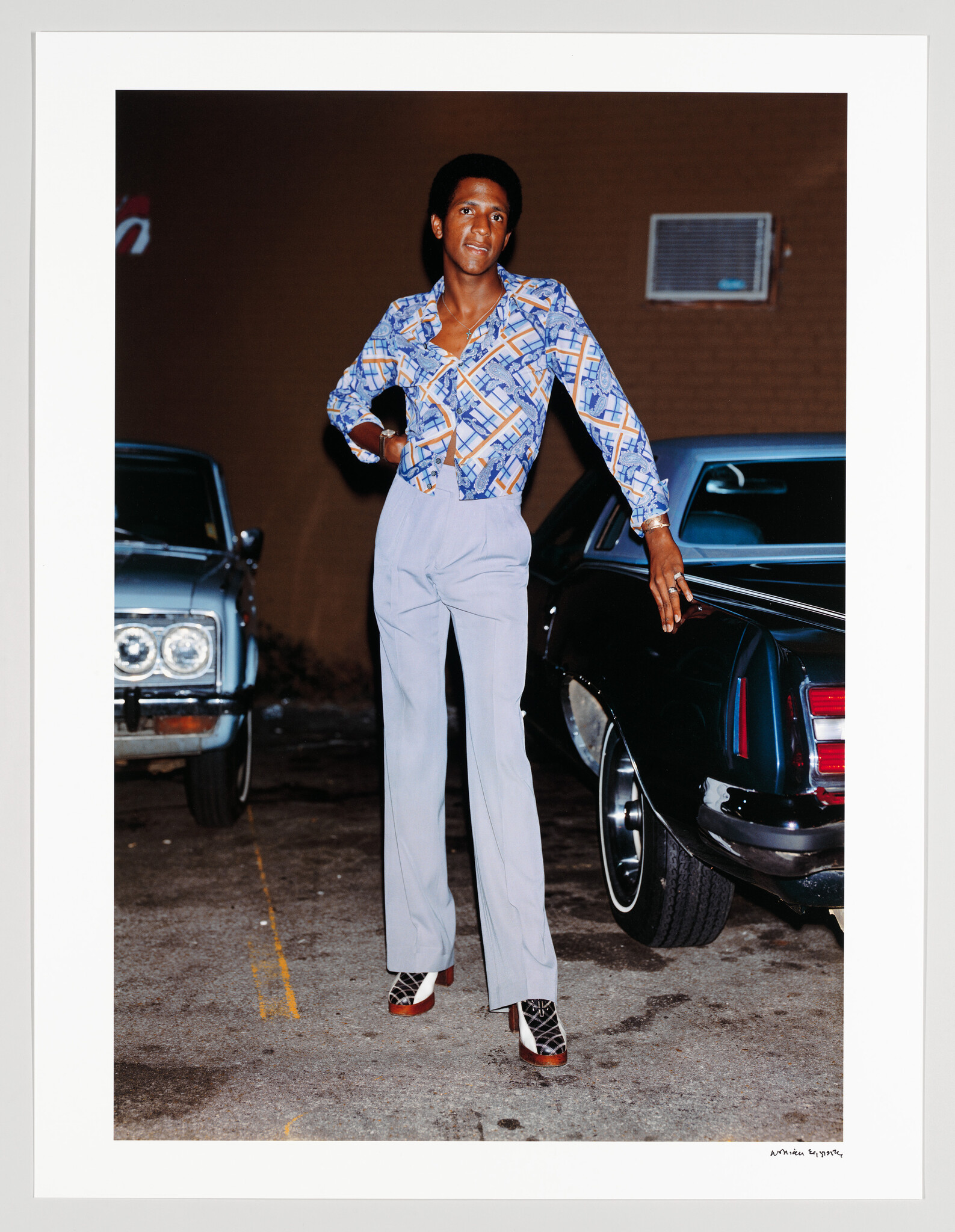 A stylish young man standing between two cars wearing a patterned shirt, high-waisted pants, and platform shoes.