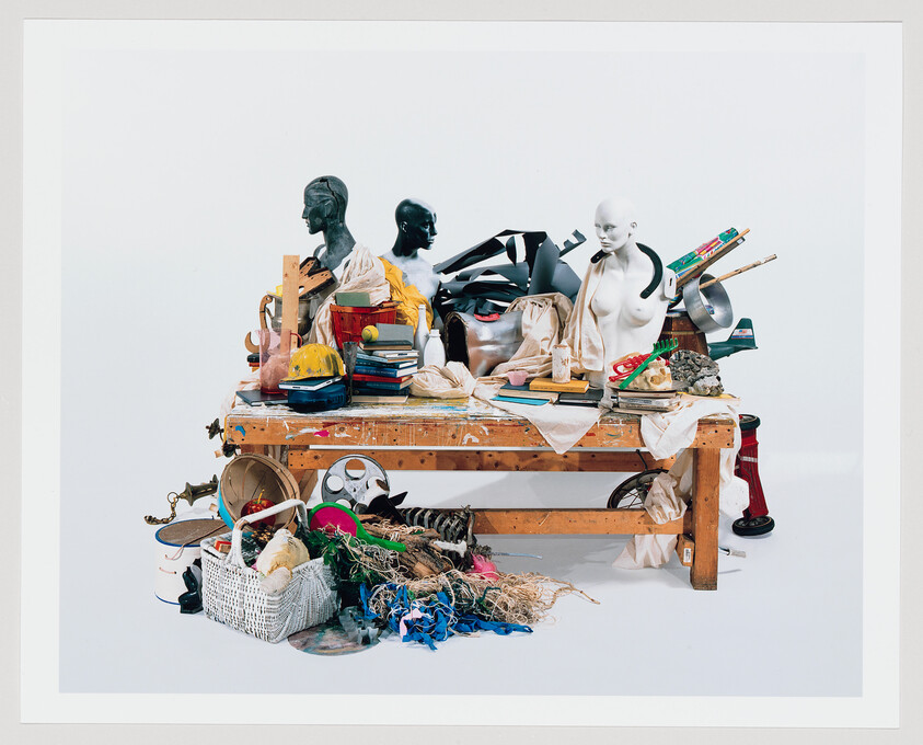 A cluttered wooden workbench holds mannequin torsos, tools, books, and assorted craft materials piled around.