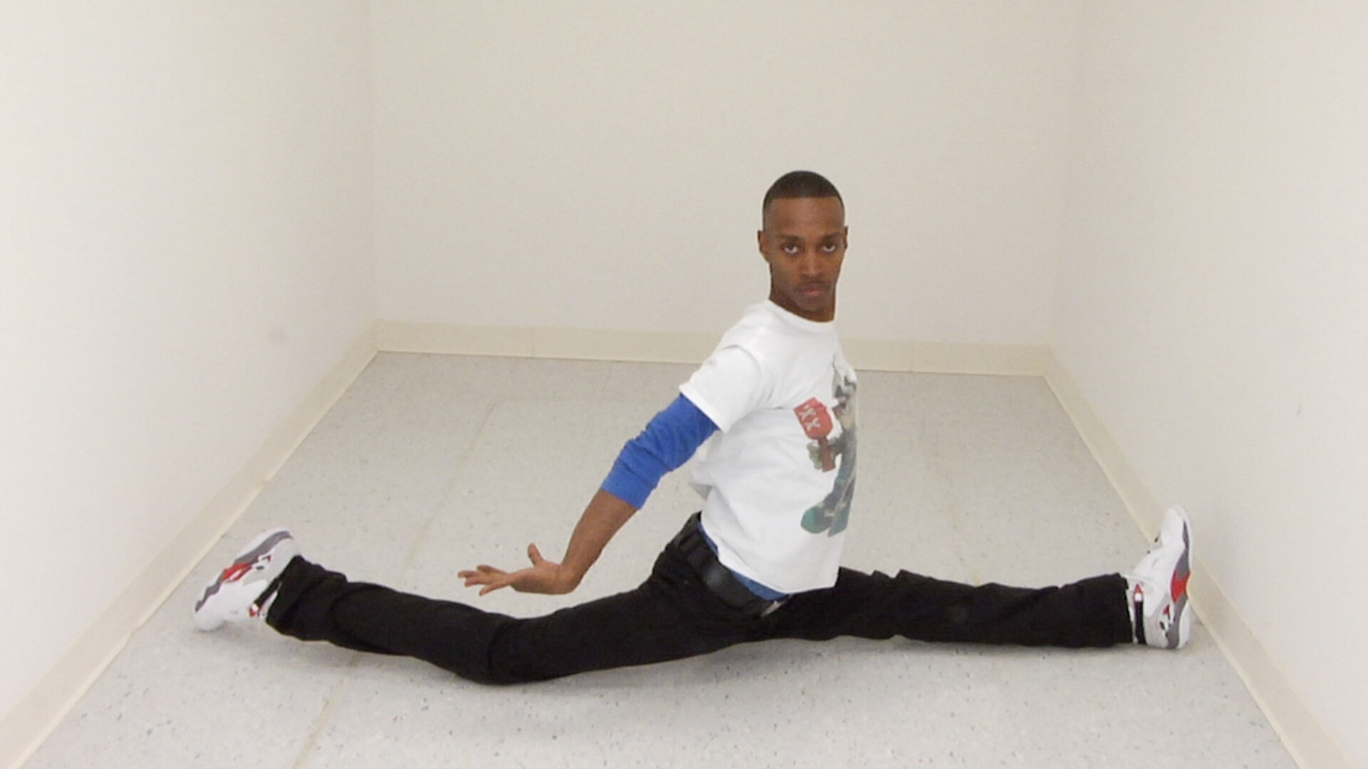 A man performs a full split on the floor between two walls, looking toward the camera.