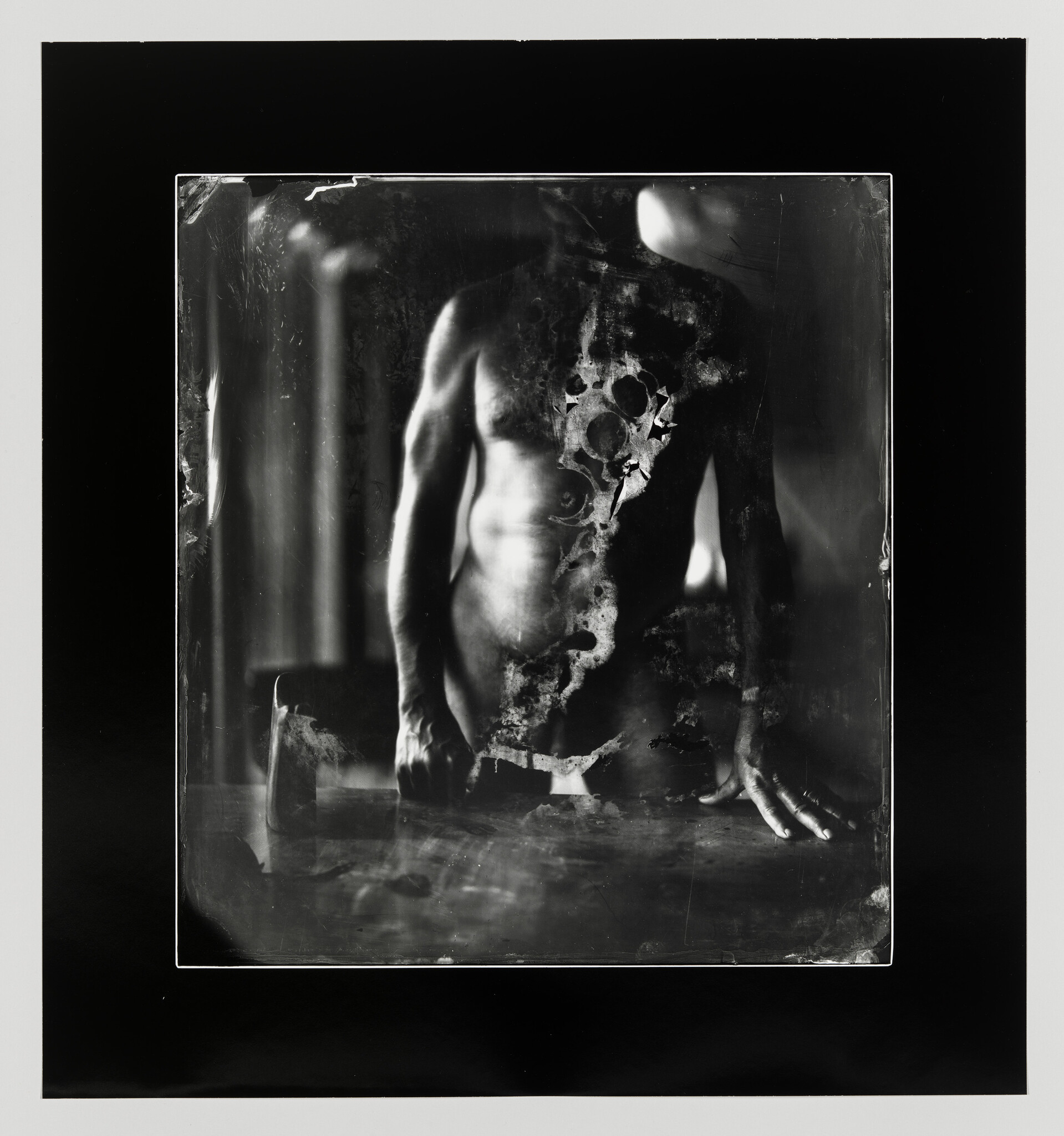 A black and white photograph depicting a damaged and deteriorated image of a human figure, with areas of the photo appearing corroded or eaten away, creating an abstract and haunting visual effect. The figure's torso and limbs are visible, but the head and parts of the body are obscured by the damage to the photograph. The edges of the photo are sharp, suggesting it is a photo of a photo, with the damaged print lying on a dark surface.