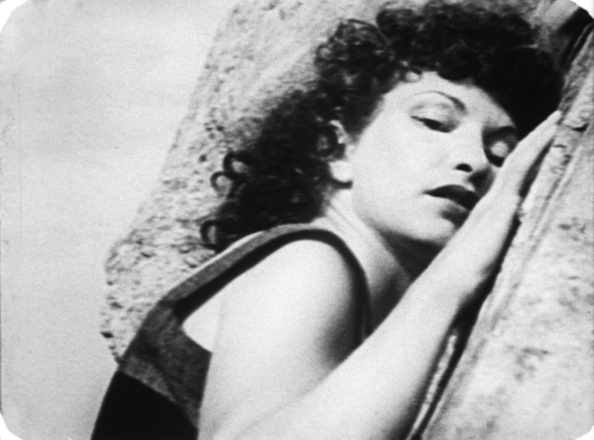 Person with curly hair leans against a large rock, looking towards the camera.