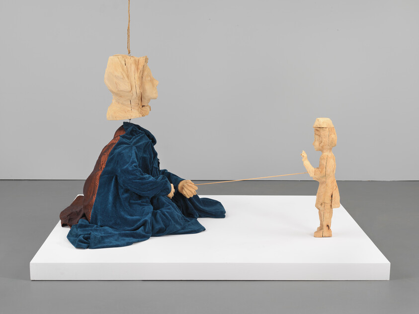 A seated wooden figure in a blue robe holds a string connecting to a small standing wooden child.