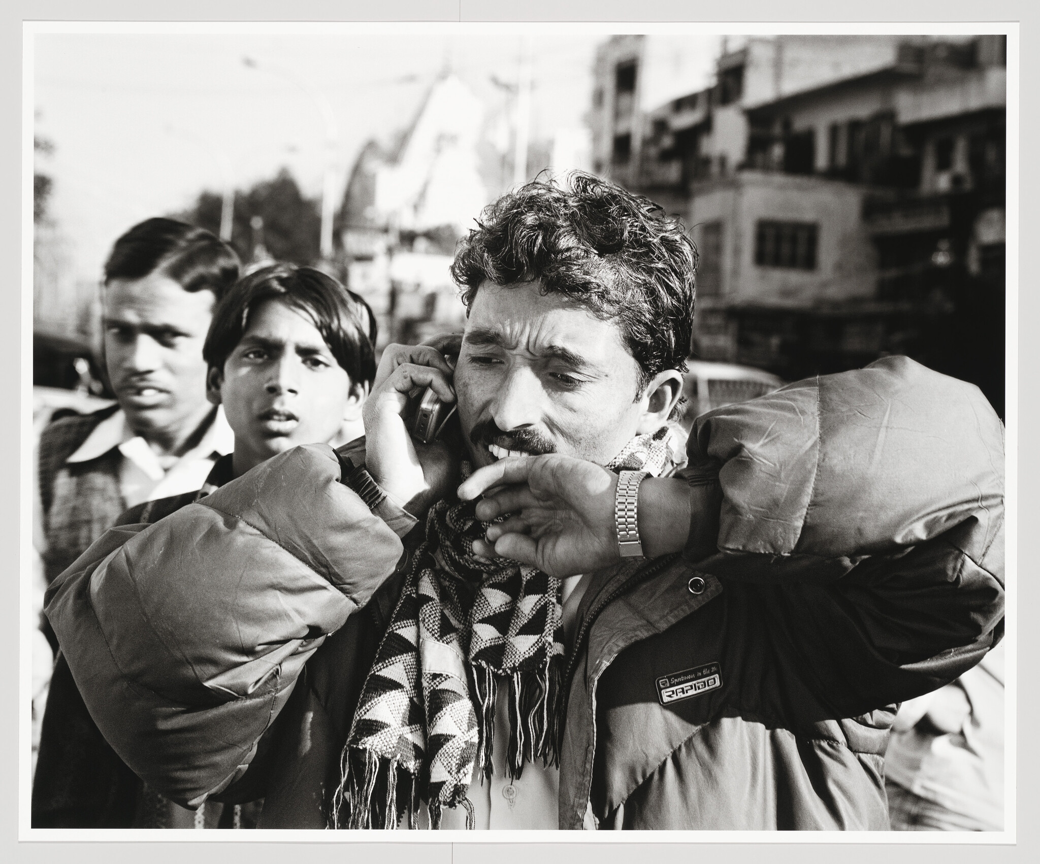 A black and white photograph capturing a moment on a busy street where a man in the foreground is talking on a mobile phone while holding a cigarette between his lips. He is wearing a jacket and a patterned scarf. Behind him, two other men appear to be looking on with curiosity. The background is slightly blurred, emphasizing the bustling urban environment.