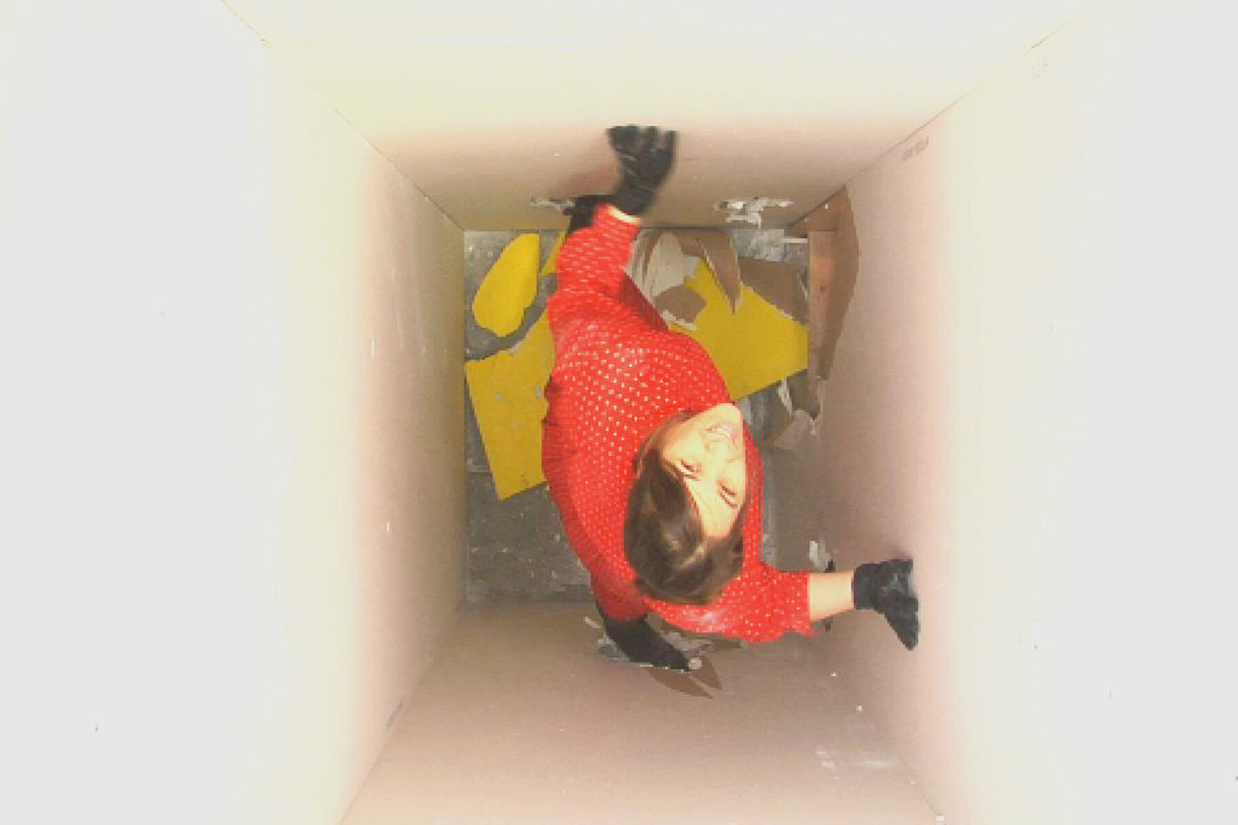 A person in a red polka dot shirt and black gloves appears to be hanging upside down in a narrow corridor with a broken wall revealing yellow insulation material. The image is rotated, creating an illusion of defying gravity.
