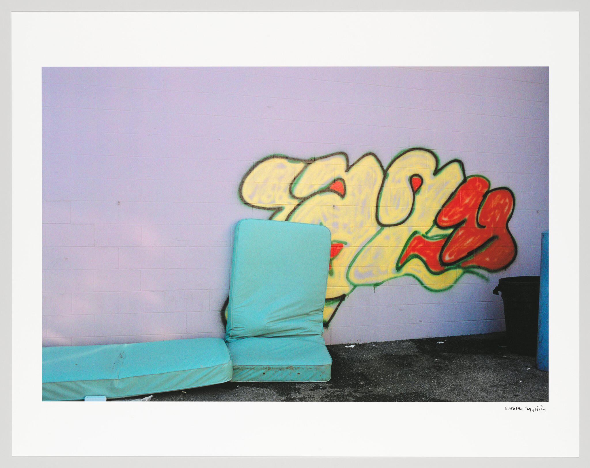 A photograph of a discarded blue mattress leaning against a wall with colorful graffiti. There's a black trash bin to the right, and the image has a white border with handwriting at the bottom right corner.