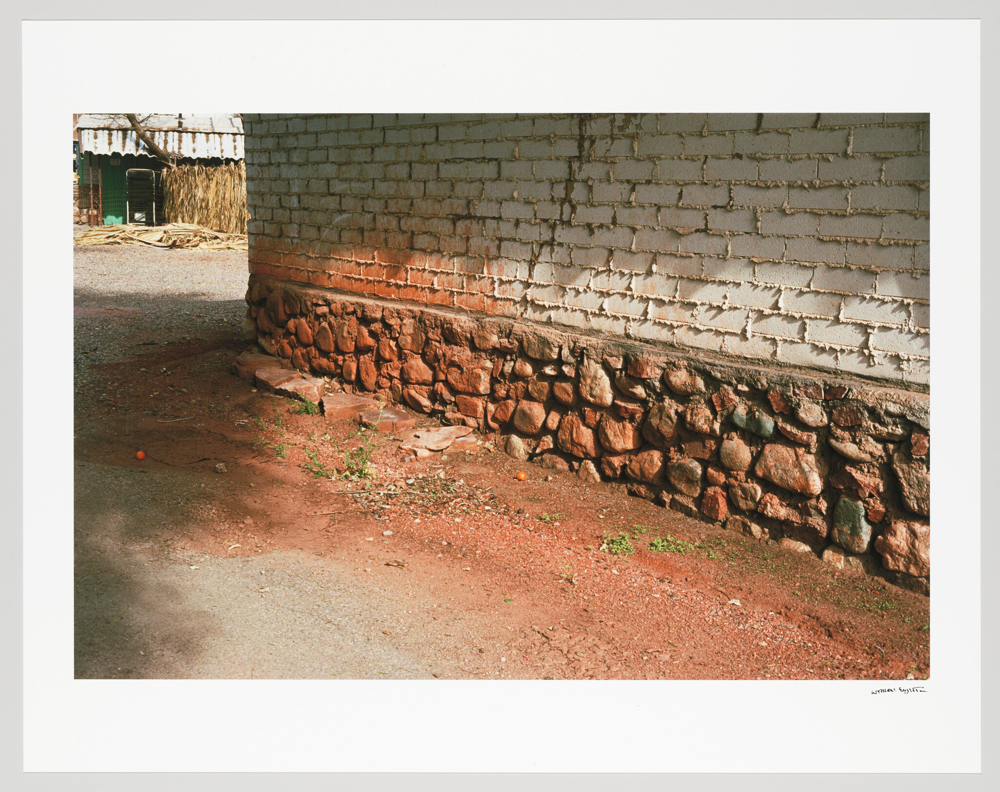 White brick wall with a stone foundation beside a dusty red dirt path with scattered debris.