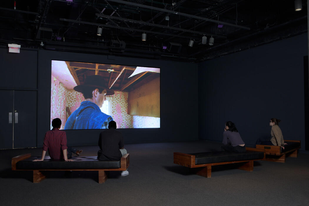 Four people sit on benches watching a large projected film of a man in a hat.