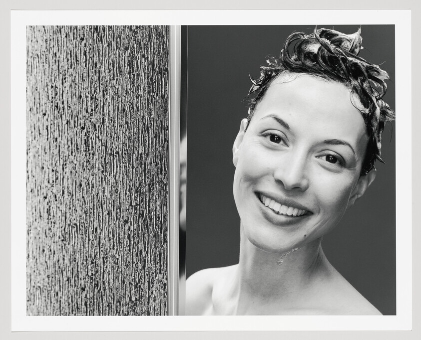 Woman with wet, lathered hair smiles while standing beside a textured shower glass.