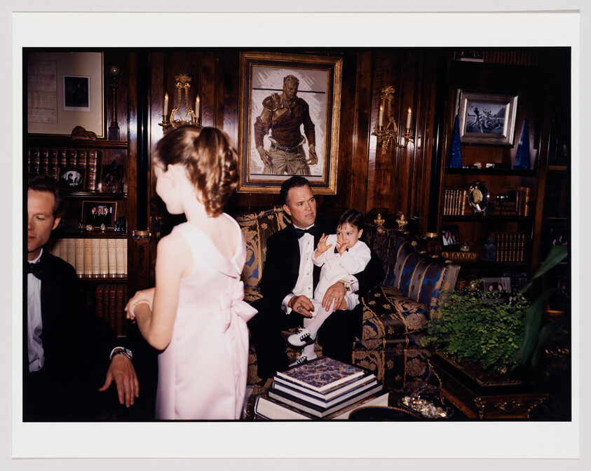 Man in tuxedo sits on couch holding young child while girl in pink dress stands nearby.