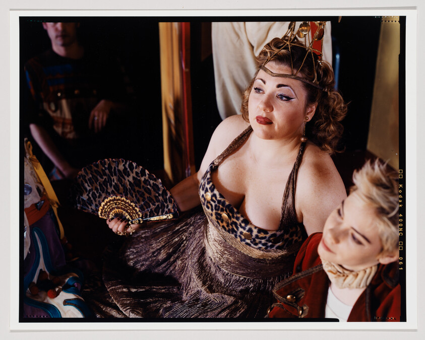 A woman in a leopard print dress and a crown sits holding a fan, with a contemplative expression. In the foreground, a person with short blonde hair and a red jacket looks down. A third individual is partially visible in the background. The setting appears to be a cluttered dressing room with various items scattered around.