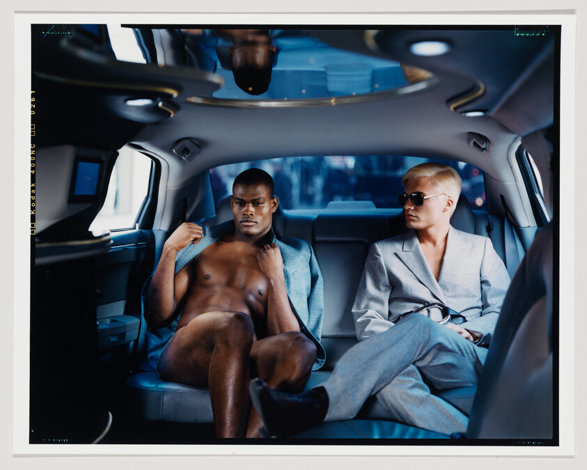 A photograph of two people sitting inside a luxury vehicle. On the left, a shirtless man with a muscular build looks directly at the camera, while on the right, a person with short blonde hair, wearing sunglasses and a gray suit, looks away. The interior of the car is spacious and modern, with blue upholstery.