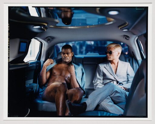 Two men sit in a car's backseat, one shirtless with a jacket and one in a suit.