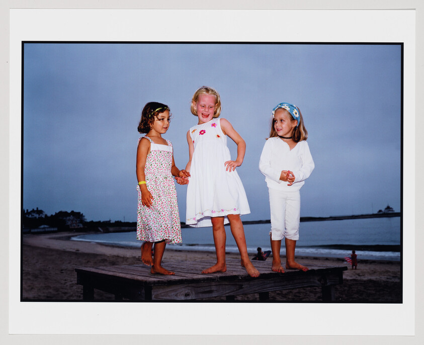 Three young girls stand barefoot on a wooden platform at the beach, smiling and holding hands.
