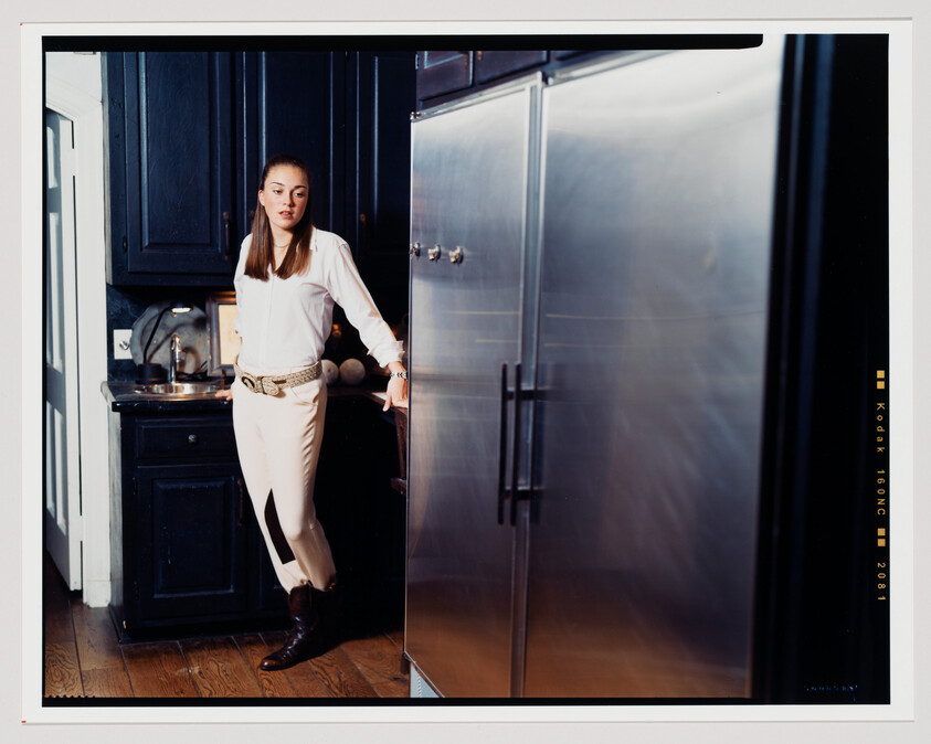 A young woman stands in a kitchen with dark cabinetry and a stainless steel refrigerator. She is dressed in a white blouse, beige pants, and brown boots, accessorized with a distinctive belt. Her pose is casual, with one hand resting on the kitchen counter and the other on the refrigerator door. The image has a film border with Kodak branding and exposure information, indicating it was taken with analog film.