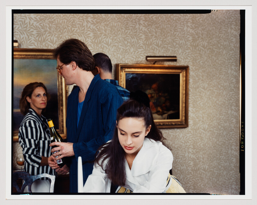 Young woman leans forward at a table while guests chat and a man holds a wine bottle.