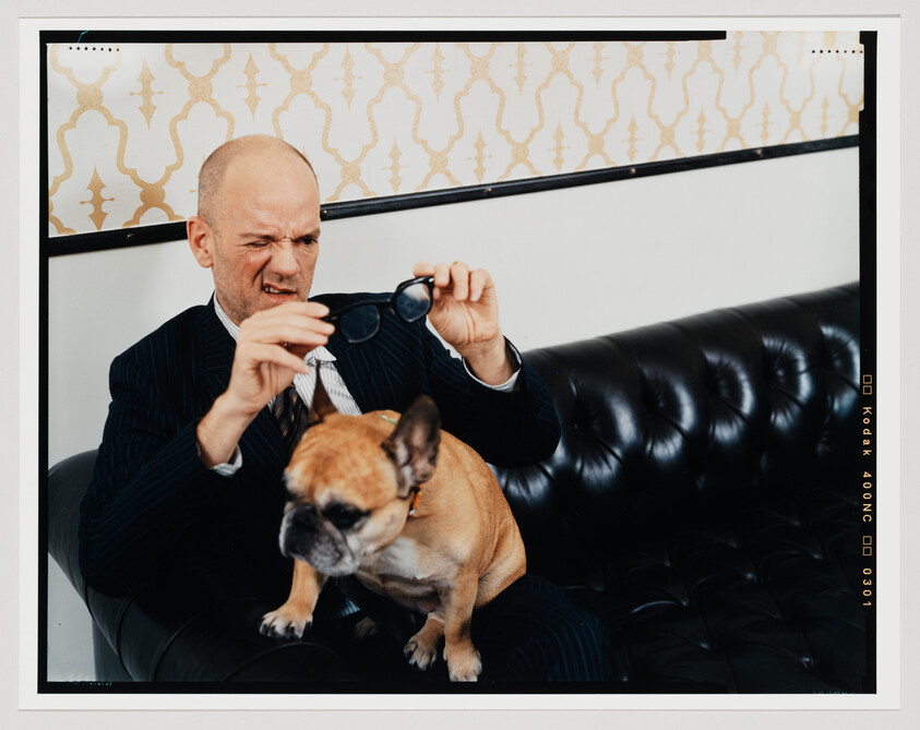 A man in a pinstripe suit sits on a black leather couch, looking quizzically at a pair of sunglasses he holds in his hands. A French bulldog sits beside him on the couch, looking off to the side. Behind them is a decorative wall with a framed picture featuring a geometric pattern. The image has a white border with film development markings, indicating it is a photograph.